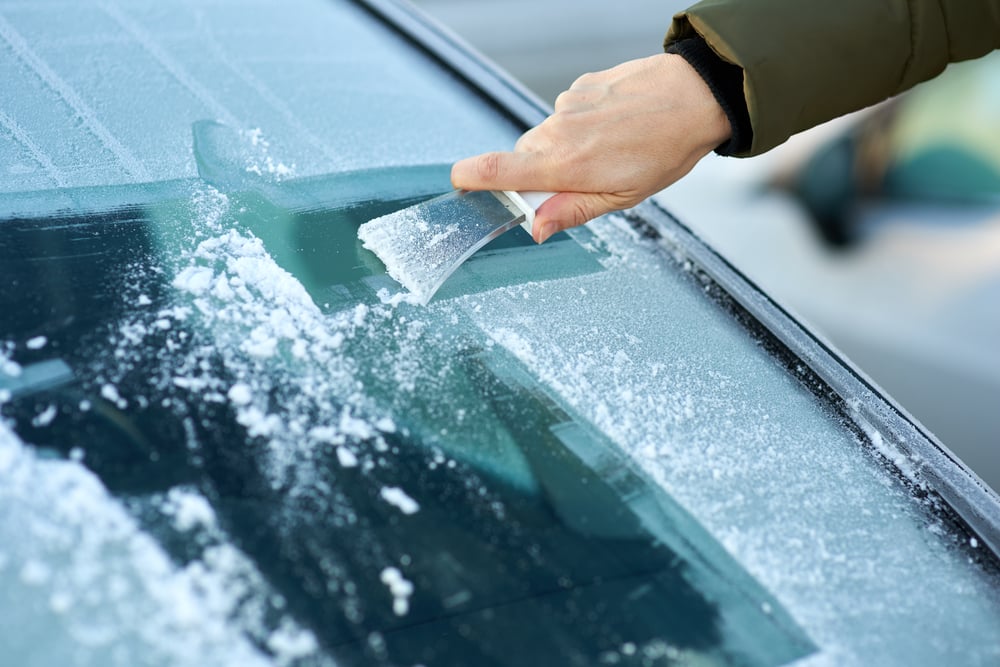 close up of ungloved man’s hand scraping ice off of windshield, green puffer coat sleeve showing